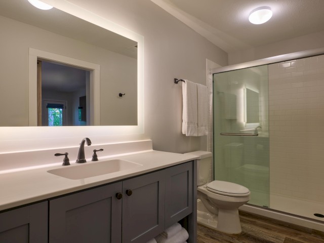 A modern bathroom with a large mirror, sink, toilet, and a glass-enclosed shower with a towel rack, and wood-look flooring.
