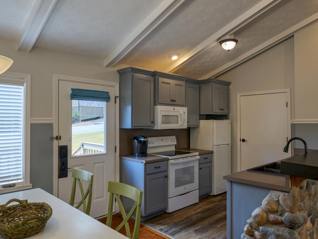This image shows a cozy kitchen with gray cabinets, a white stove and microwave, a small dining area with green chairs, and stone accents.
