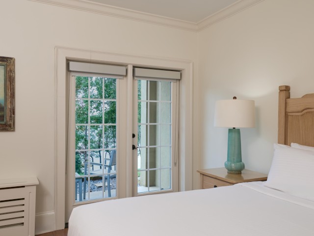 A bright bedroom features a bed, a nightstand with a lamp, a painting, and French doors leading to a balcony with outdoor seating.