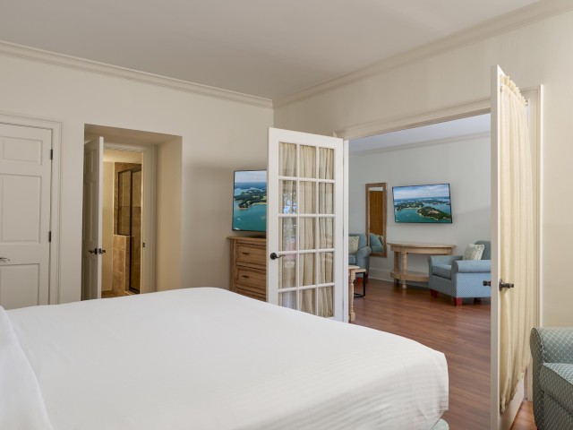 The image shows a bedroom with a white bed, connecting to a sitting area with armchairs, a dresser, and a TV through French doors.