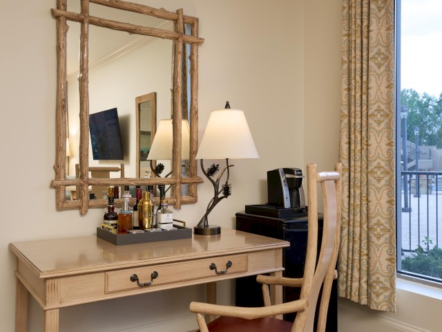 A desk with a mirror, lamp, coffee maker, and a tray of bottles is near a window with curtains overlooking an outdoor view.