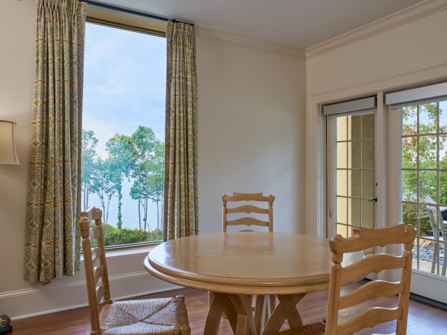 A dining area with a round wooden table and chairs, large window with a nature view, curtains, lamp, and doorway to a patio outside.