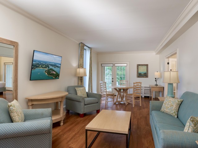 The image shows a cozy living room with sofas, armchairs, a coffee table, a TV, and a dining area near large windows.
