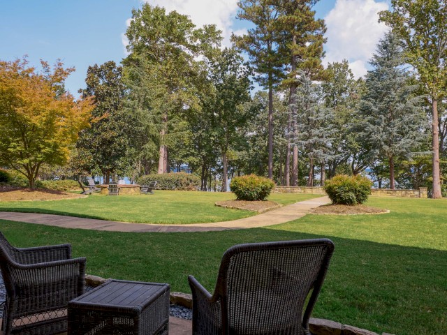 Two chairs and a table on a patio overlook a grassy area with trees and a path under a blue sky.