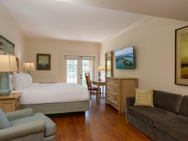 A cozy hotel room with a bed, TV, sofa, chairs, and artwork. Warm lighting and wood floors create a comfortable atmosphere.