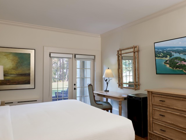 A cozy bedroom with a view of the outdoors, featuring a bed, desk, mirror, TV, and natural light from double doors, creating a relaxing atmosphere.