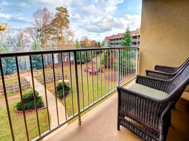 The image shows a balcony with two wicker chairs overlooking a landscaped garden area, trees, and nearby buildings under a partly cloudy sky.