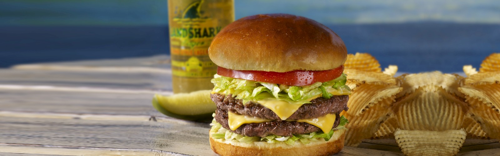 A double cheeseburger with lettuce and tomato, a pile of potato chips, a pickle, and a bottle in the background.