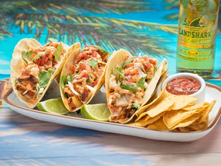 Fish tacos with lime, a side of chips and salsa, and a bottle of LandShark Lager on a colorful beach-themed background.
