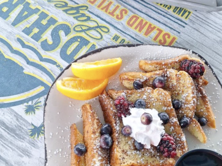 French toast with berries, whipped cream, and syrup on a plate with orange slices, and a tray of fries in the background.