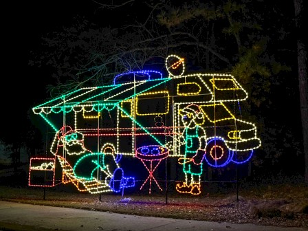 A colorful illuminated bus sculpture at night, with people lounging, a table and cooler, glowing outlines in green, yellow, blue, and red.