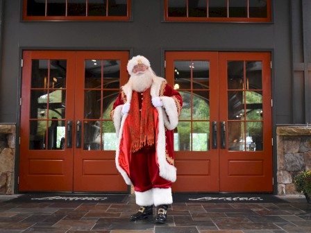 A person dressed as Santa Claus stands in front of large red doors, wearing a red suit with white trim.