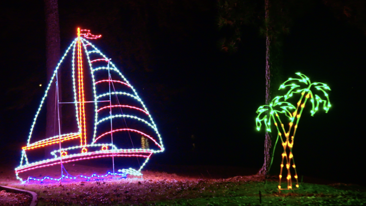 A night scene with illuminated decorations: a sailboat outlined in multicolor lights on the left and a glowing palm tree on the right, set on a dark yard.