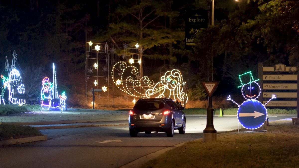 A car approaches a holiday light display featuring illuminated figures and decorations, including a snowman and swirling shapes, along a road.