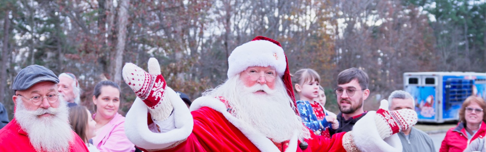 A cheerful Santa Claus waves to a crowd in a park, surrounded by families and kids, as winter trees frame the scene.