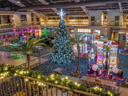 A decorated indoor arcade with a Christmas tree, palm trees, and colorful arcade games and rides, lit with festive lights and ornaments.