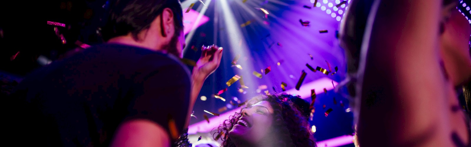 People are dancing and enjoying a nightclub party under colorful lights with confetti falling around them.