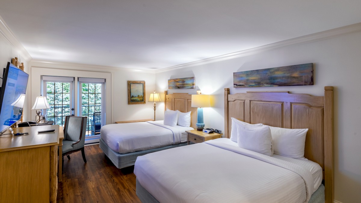 The image shows a hotel room with two beds, wooden furniture, a desk with a chair, lamps, and a large window with curtains.