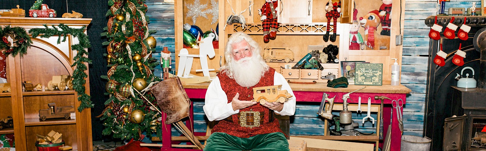 A person dressed as Santa in a workshop setting, surrounded by Christmas decorations and gifts, is working on a wooden toy.