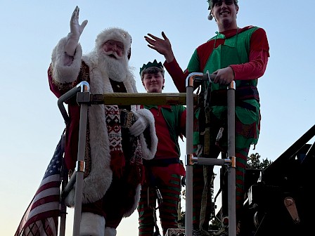 Santa Claus and two people dressed as elves waving from a fire truck with an American flag.