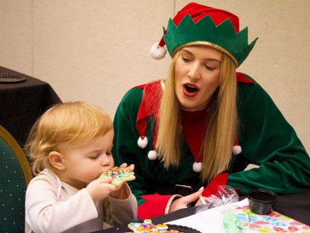 A child enjoys a decorated cookie while sitting next to a person dressed as a festive elf at a table with supplies and decorations.