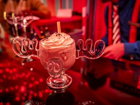 A drink served in a glass shaped like a moose head, topped with cream, sits on a bar counter under red lighting.