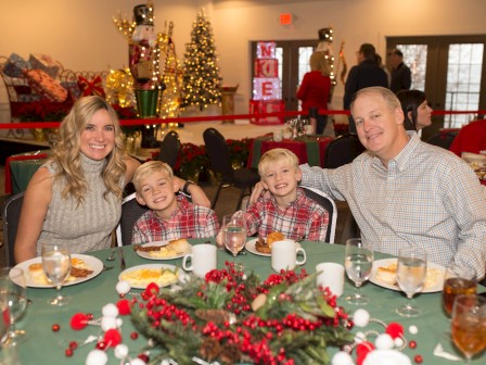 A family of four sits at a festive holiday table with greenery and berries, smiling for the camera in a decorated banquet hall.