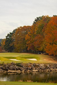 A scenic golf course by a calm lake, surrounded by autumn trees with vibrant foliage and a rocky shoreline.