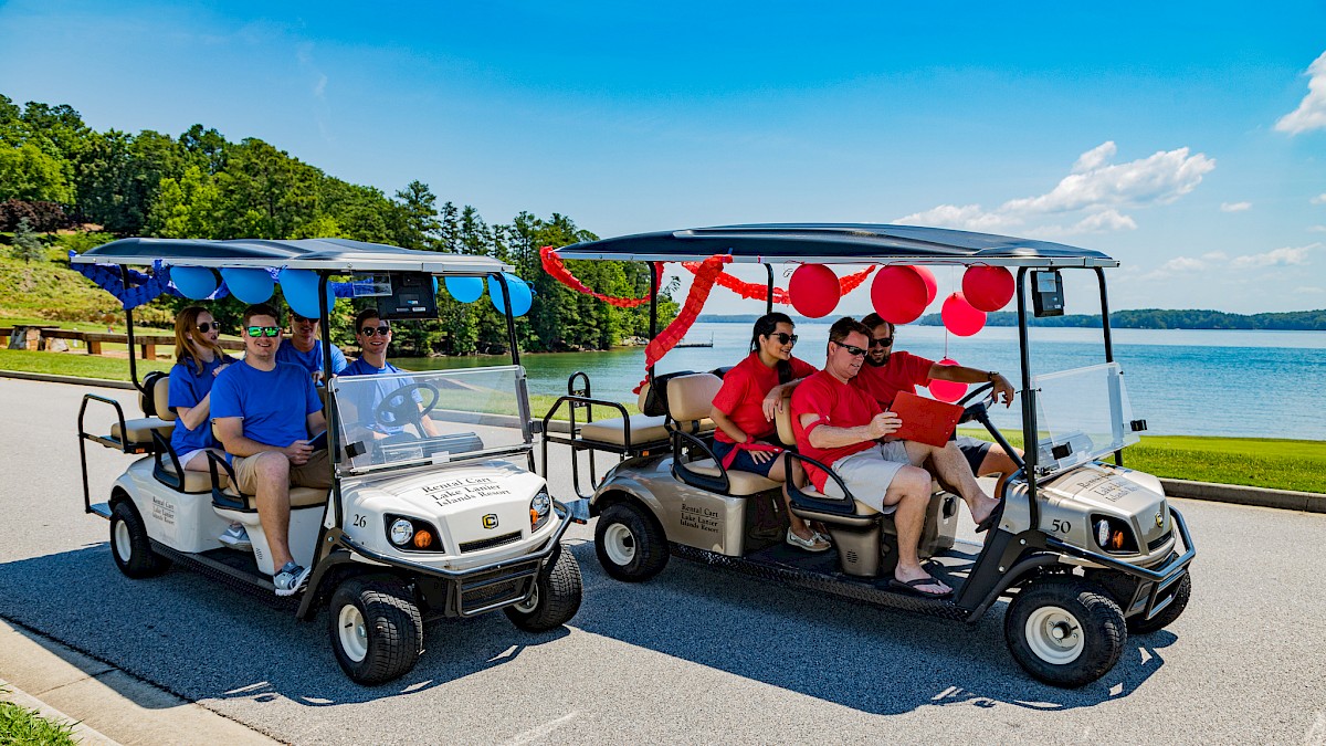 Two decorated golf carts carry groups of people near a lake, celebrating a festive occasion on a sunny day.