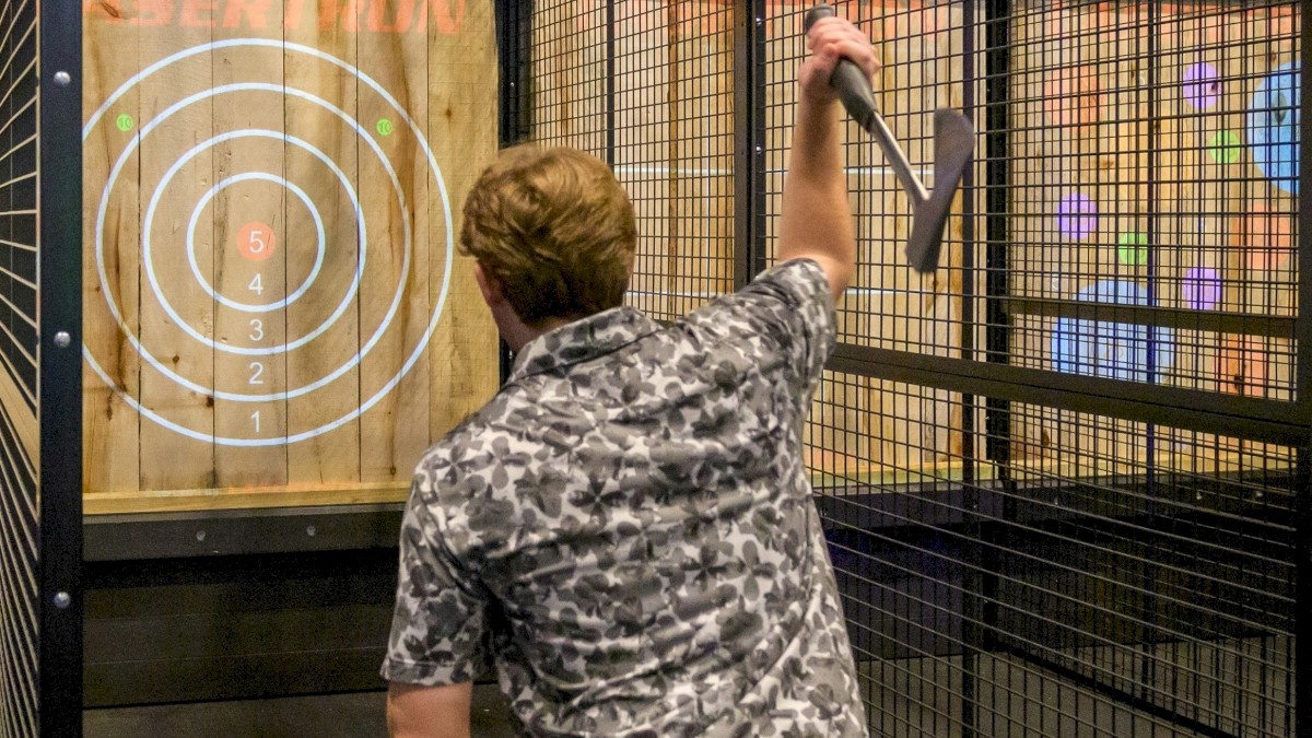 A person is throwing an axe at a circular target in a fenced area, wearing a patterned shirt and light pants.