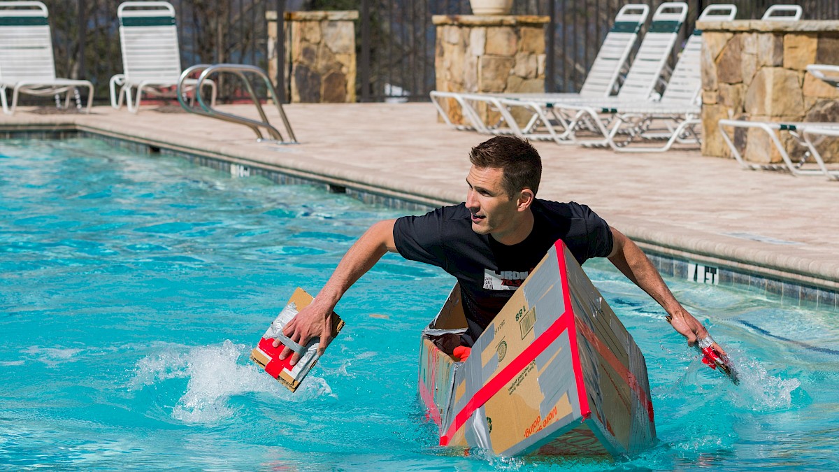 A man in a black shirt is in a pool, holding a cardboard box, with lounge chairs and stone pillars in the background.