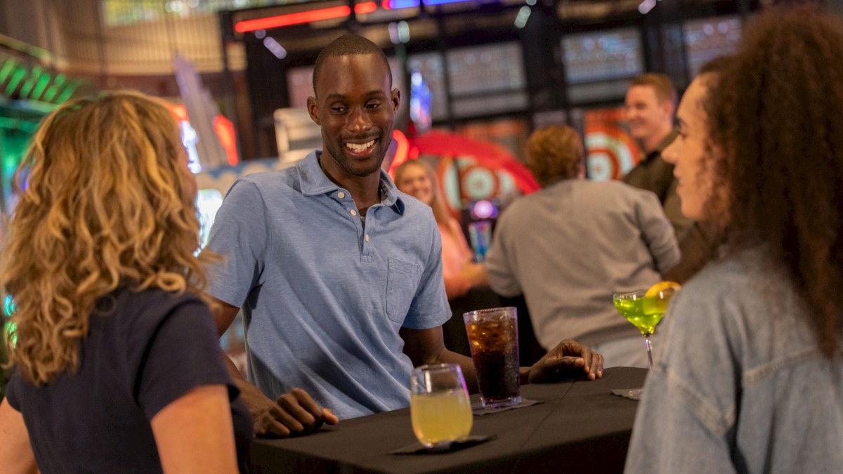 People are socializing at a table in a lively venue, holding drinks and smiling, with colorful lights in the background.