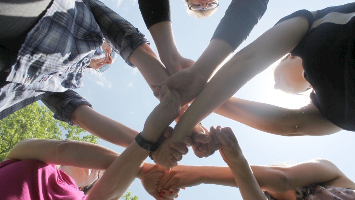 A group of people are outdoors, reaching out and stacking their hands together in a teamwork gesture against a blue sky.