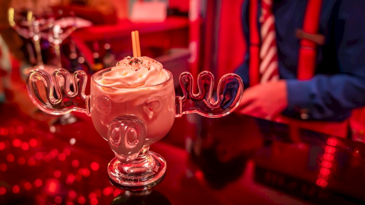 A whimsical moose-shaped mug contains a creamy drink, topped with whipped cream. A person in colorful attire is in the background.