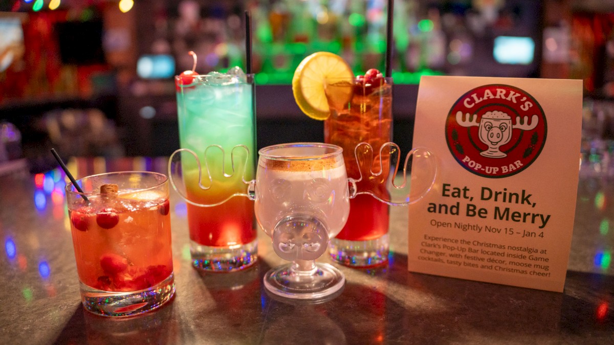 A row of colorful cocktails on a bar counter with ice, fruit garnishes, and a Clark’s U bar sign promoting “Eat, Drink, and Be Merry.”
