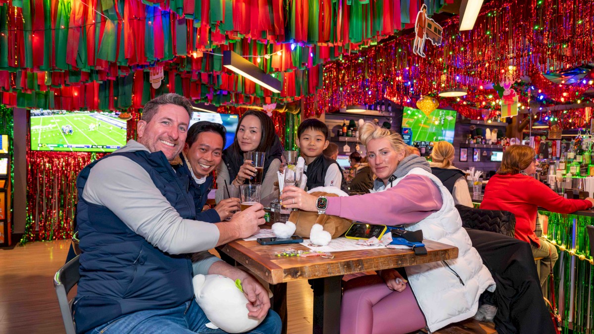A group of friends at a colorful, festive restaurant, sitting around a wooden table, toasting drinks, with vibrant hanging streamers all around.