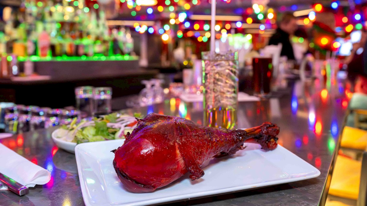 Crispy roasted duck on a white plate, set on a shiny bar counter with colorful neon lights and a busy, festive diner vibe.