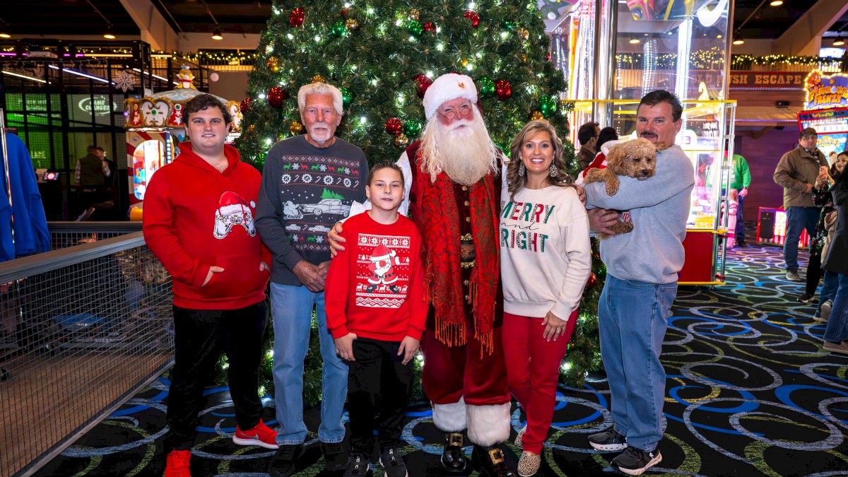 A festive group pose with Santa Claus in the center, kids and adults gathered around a decorated Christmas tree in a bright arcade setting.