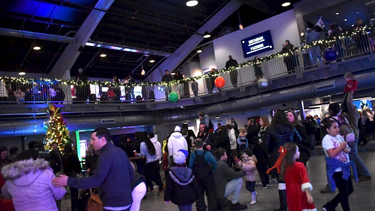 A festive indoors event with people gathered, Christmas tree, decorations, and children in holiday outfits seen.