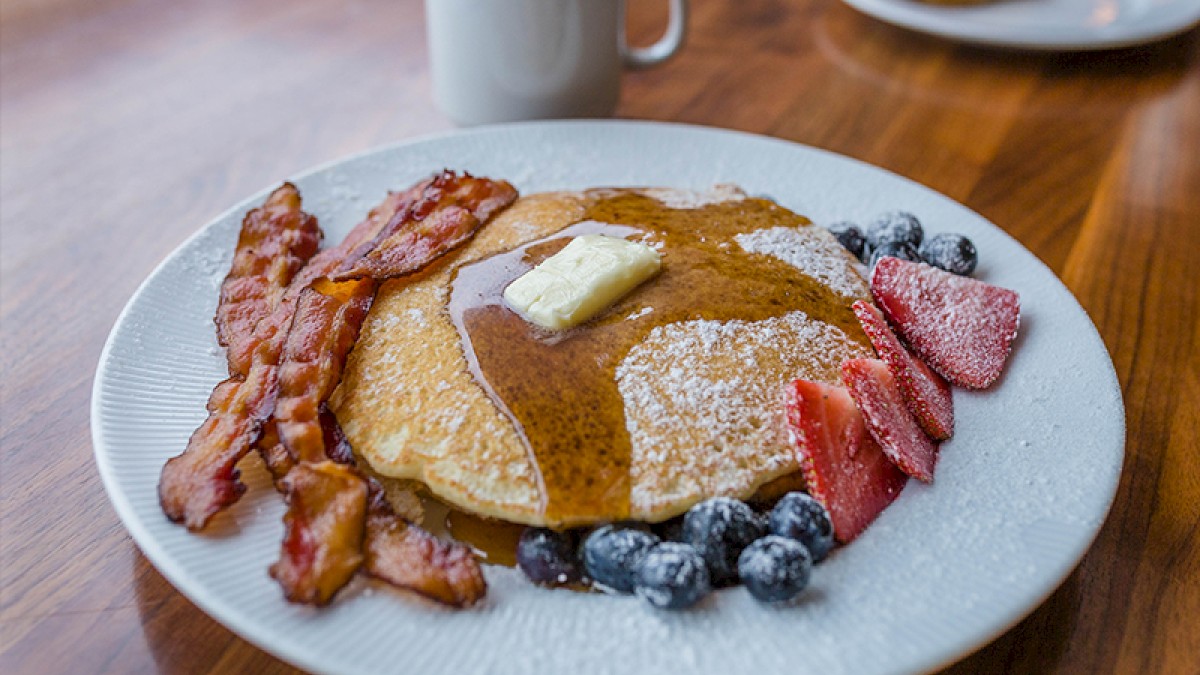 A plate of pancakes with syrup and butter, bacon, blueberries, and strawberries, accompanied by a mug on a wooden table.
