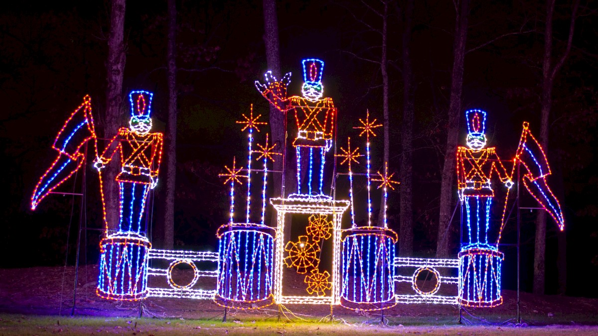 Three illuminated toy soldiers and drums are displayed in a festive, colorful arrangement, set against a dark background.