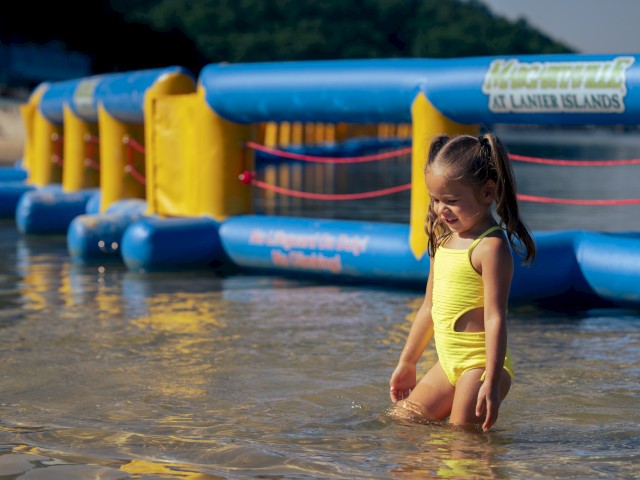 A young girl in a yellow swimsuit enjoys wading in shallow water with a colorful inflatable water obstacle course in the background.