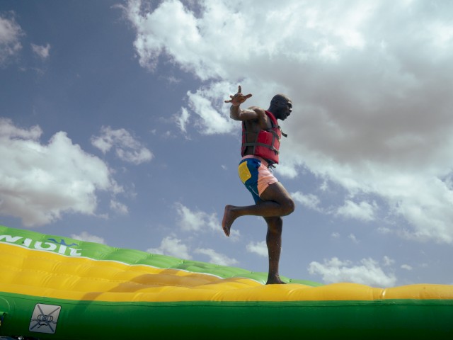 A man balances on one foot on an inflatable water slide outdoors under a partly cloudy sky.