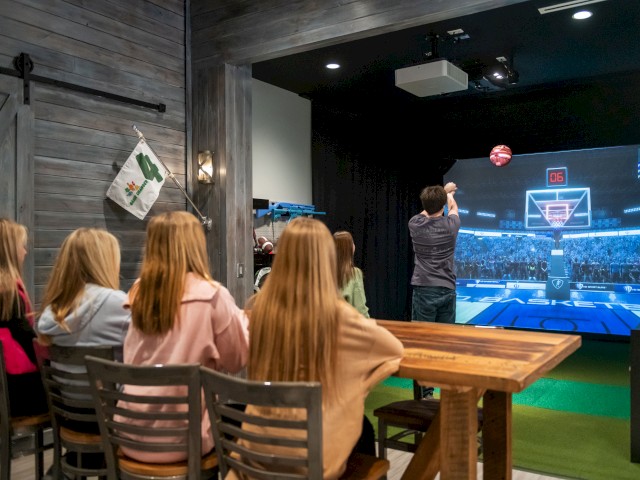 A group of young women watching a basketball game on a screen while a man is shooting the ball inside a recreational indoor space.