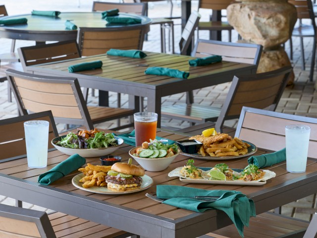 A table with various dishes, salads, fried foods, a burger, drinks, and green napkins at an outdoor restaurant.