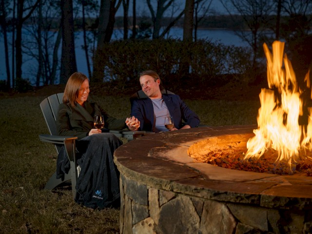 A couple is sitting near a campfire, holding hands and enjoying drinks at dusk by a lakeside.