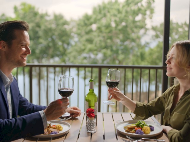 A couple enjoying a romantic dinner outside with wine, salads, and a scenic view of greenery and water.