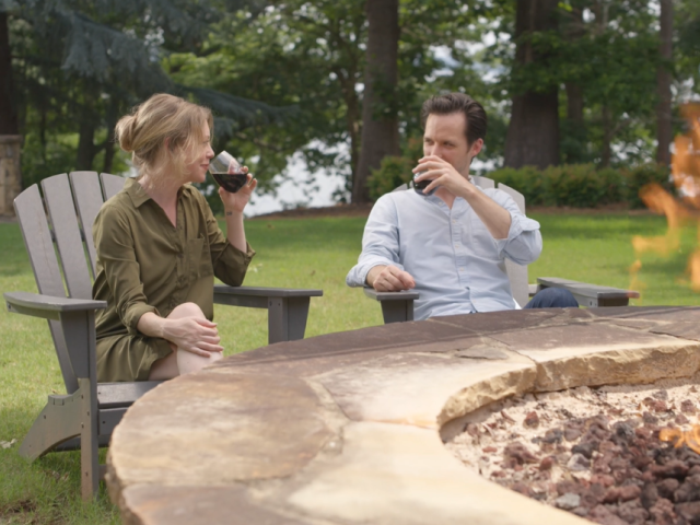 A man and woman sit near a fire pit outdoors, drinking red wine and enjoying a relaxing moment in a green, wooded backyard.