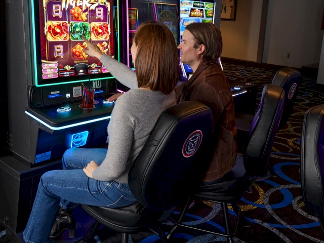 Two people are playing a slot machine game in a casino, with colorful screens and a patterned carpet visible.