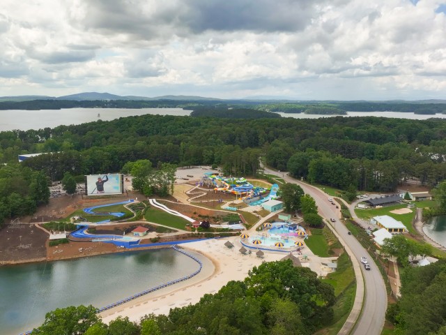 This image shows an aerial view of a water park surrounded by trees, lakes, and a road, with multi-colored water slides and a large screen.